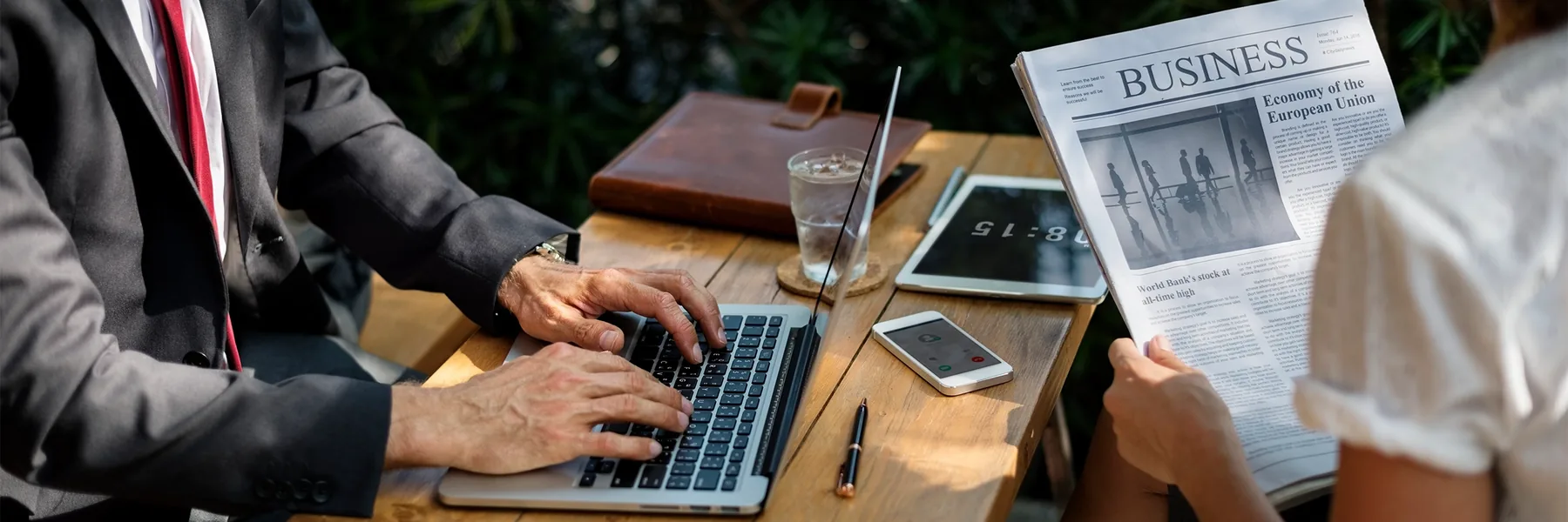 Two colleagues at an outdoor table, one typing on a laptop and one reading a business newspaper